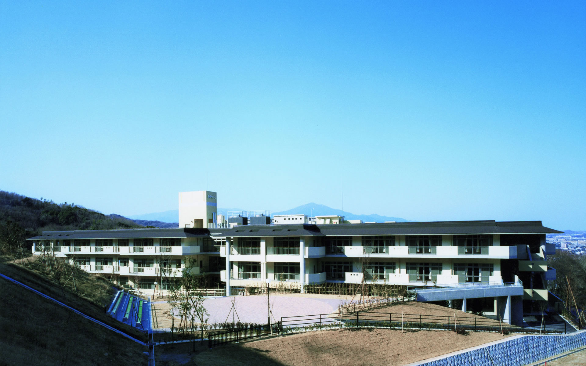 山々を背景とした介護施設の建物外観。複数階建ての白色モダン建築で、前庭に工事中の様子が見られます。